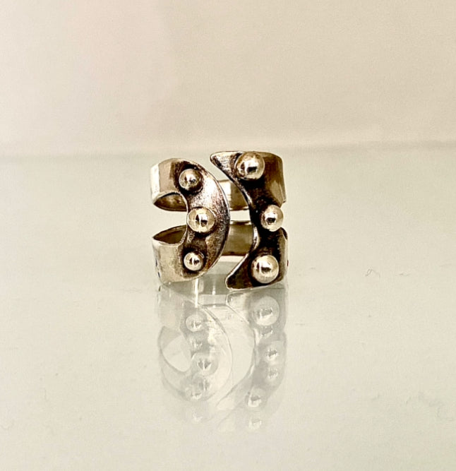 A silver-toned ring with an abstract sculptural design and raised metallic bead details, displayed on a reflective white surface against a light background.