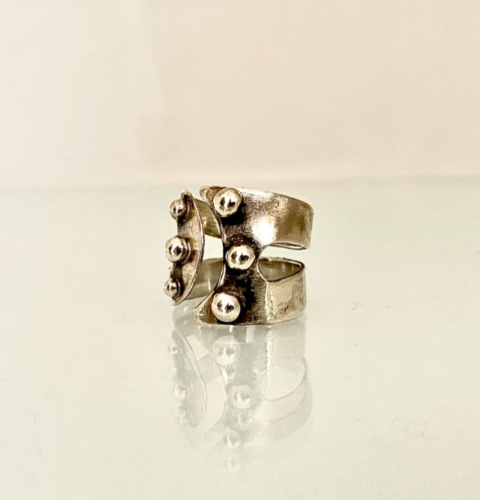 A textured silver ring with a wide band and protruding spherical bead details, displayed on a reflective white surface against a light beige background. MAYBE adjustable sterling silver ring with six hand fused granules.