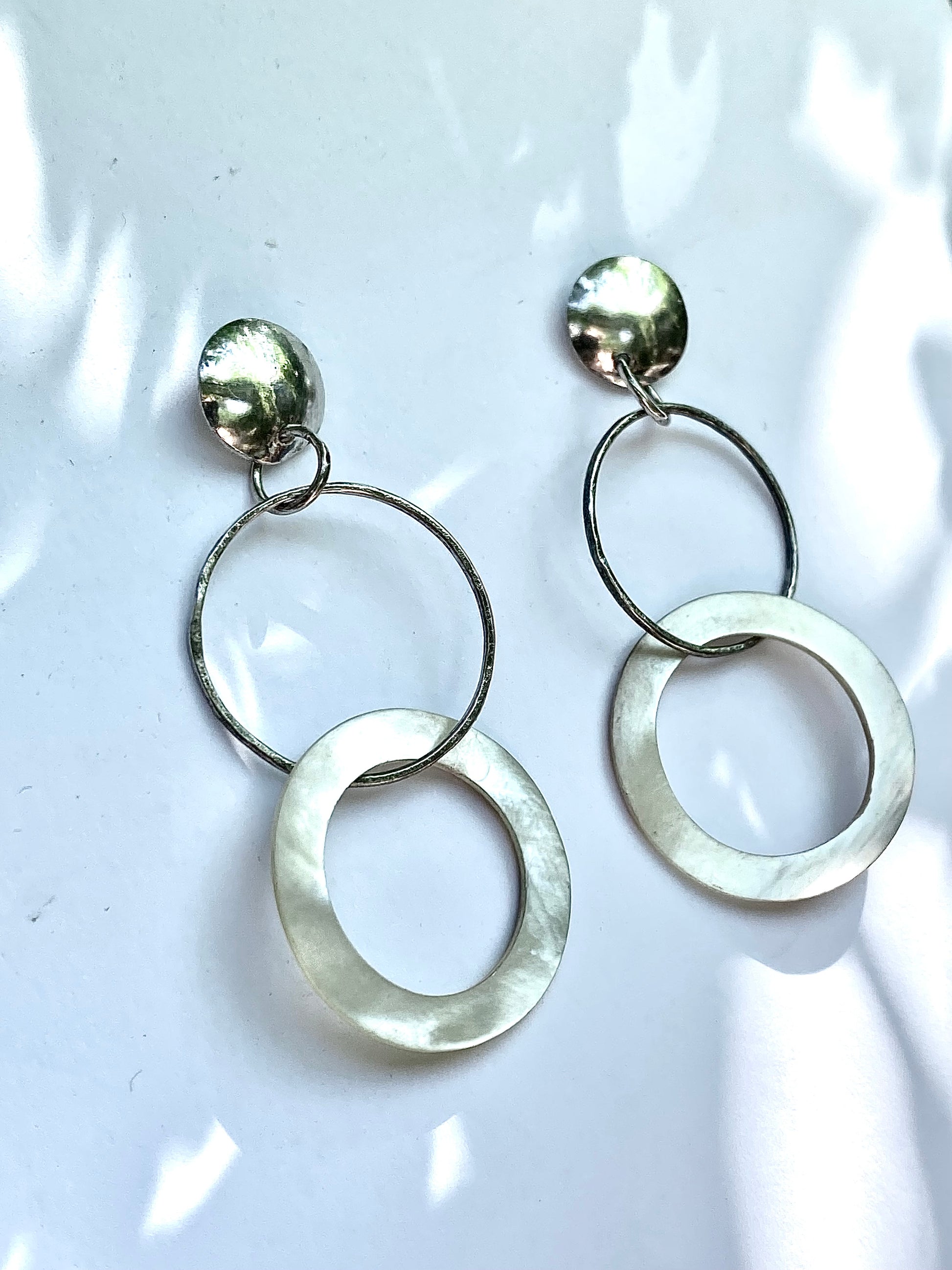 A pair of silver-toned dangle earrings with a textured circular stud, a thin hanging ring, and a larger open oval drop, displayed on a white surface with soft leaf shadows.