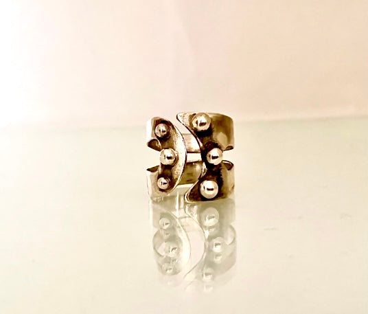 A silver-toned metal ring with a sculptural, organic shape and raised metallic bead details, displayed on a reflective white surface against a light beige background. MAYBE open band sculptural silver ring.
