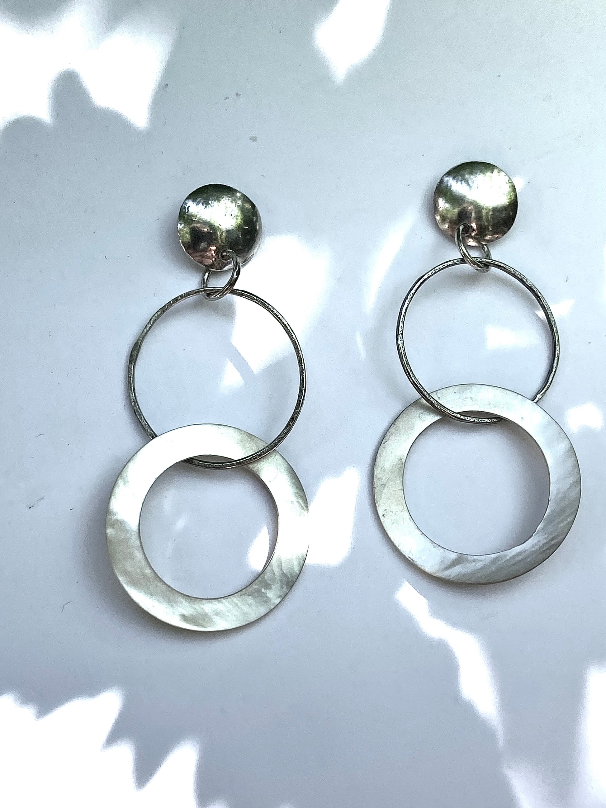 A pair of silver dangle earrings with a textured circular disc design, displayed against a white background with soft leaf shadows.