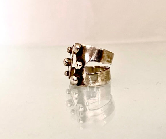 A silver-toned ring with a textured, hammered finish and small metallic bead details on the front, displayed on a reflective white surface against a light background. MAYBE open band silver ring showing tapered back for comfort, side view.