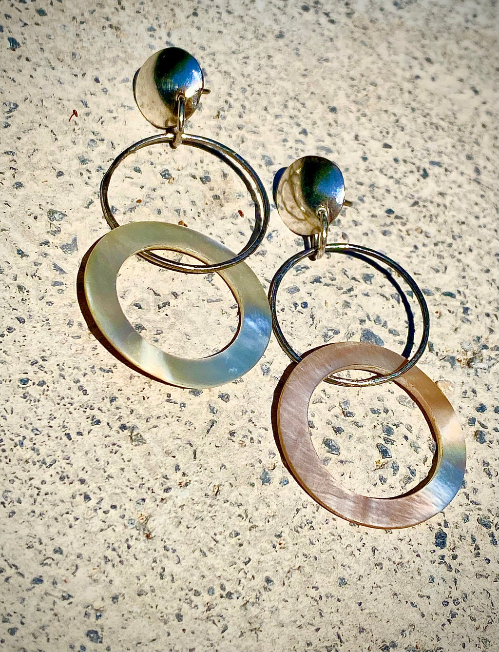 A pair of statement dangle earrings with large circular disc elements and thin interlocking rings, photographed on a textured light stone surface.
