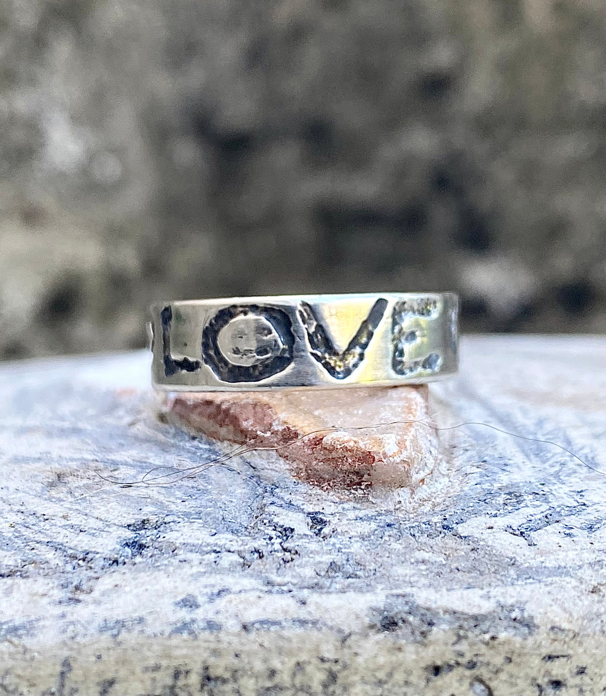 A silver ring engraved with the word 'LOVE' in raised, rustic letters, resting on a textured stone surface with a blurred natural background.